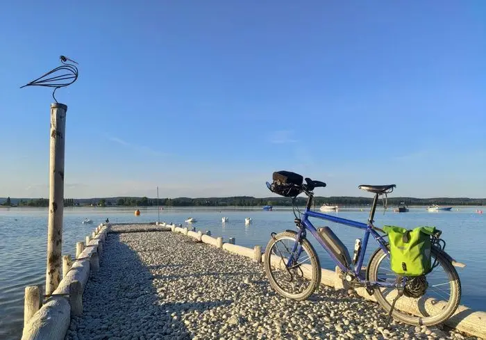 Das Bild zeigt eine ruhige, malerische Szene an einem See. Ein Fahrrad steht am Rand eines Steges voller Schotter. Auf einem Holzpfosten befindet sich eine Vogel-Skulptur. Der Himmel und das Wasser sind klar und blau. In der Ferne liegen kleinen Boote.
