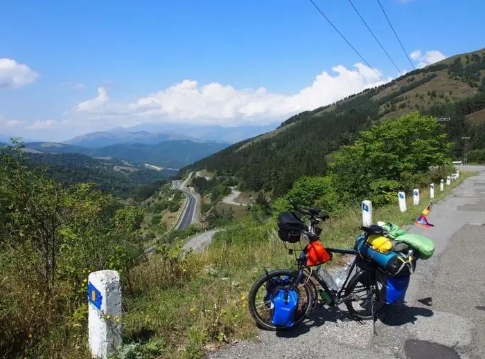 Das Bild zeigt ein vollgepacktes Tourenrad, am Rande einer Straße in einer bergigen Landschaft. Die Straße schlängelt sich durch grüne Hügel und Berge unter einem klaren blauen Himmel.