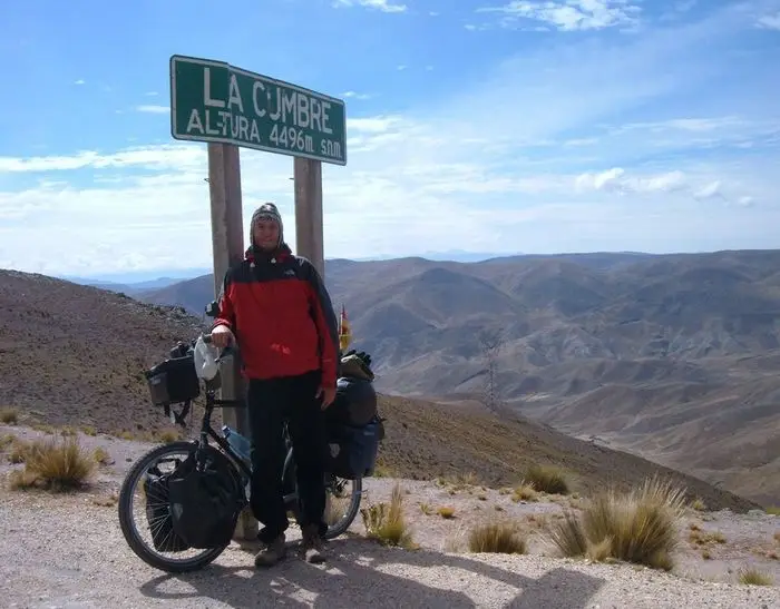 Das Bild zeigt eine Person mit einem vollgepackten Tourenrad neben einem Schild, auf dem “LA CUMBRE ALTURA 4496m s.n.m.” steht. Die Person und das Fahrrad sind vor einer beeindruckenden Berglandschaft positioniert.
