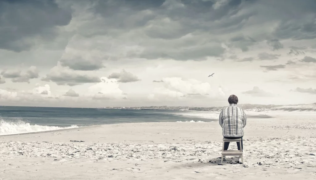 Das Bild zeigt einen ruhigen und nachdenklichen Moment an einem Strand. Eine Frau sitzt auf einem Stuhl und blickt über den Ozean. Der Himmel ist von dramatischen Wolken dominiert. Das Bild veranschaulicht einen einsamen Moment der Reflexion an einem leeren Strand unter einem ausdrucksstarken, bewölkten Himmel und ruft Gefühle von Einsamkeit und Selbstbetrachtung hervor.