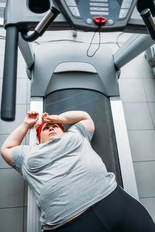Das Bild zeigt eine übergewichtige Person, die nach einem möglicherweise intensiven Training, völlig erschöpft auf einem Laufband in einem Fitnessstudio liegt und verdeutlicht die körperliche Anstrengung beim Sporttreiben.