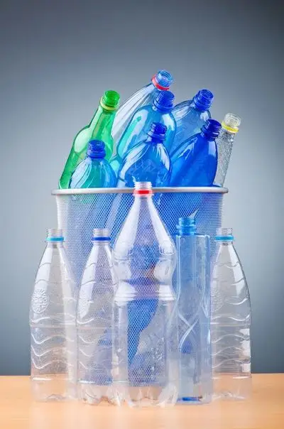 Das Bild zeigt eine Sammlung von farbigen und transparenten Plastikflaschen, die aus einem metallischen Drahtkorb für Abfälle herausquellen und veranschaulichen Themen wie Recycling oder Abfallmanagement.
