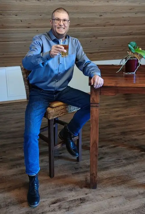 Das Bild zeigt eine Person, die in einer ästhetisch ansprechenden Holzumgebung an einem Holztisch sitzt und ein Getränk in einem Glas geniesst.
