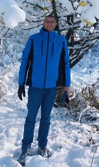 Das Bild zeigt Patrick, der in einer verschneiten Landschaft steht. Im Hintergrund steht ein schneebedeckter Baum mit Äpfeln. Das Bild vermittelt eine ruhige und kalte Atmosphäre und zeigt die Widerstandsfähigkeit und den Kontrast der Natur selbst unter widrigen Wetterbedingungen.