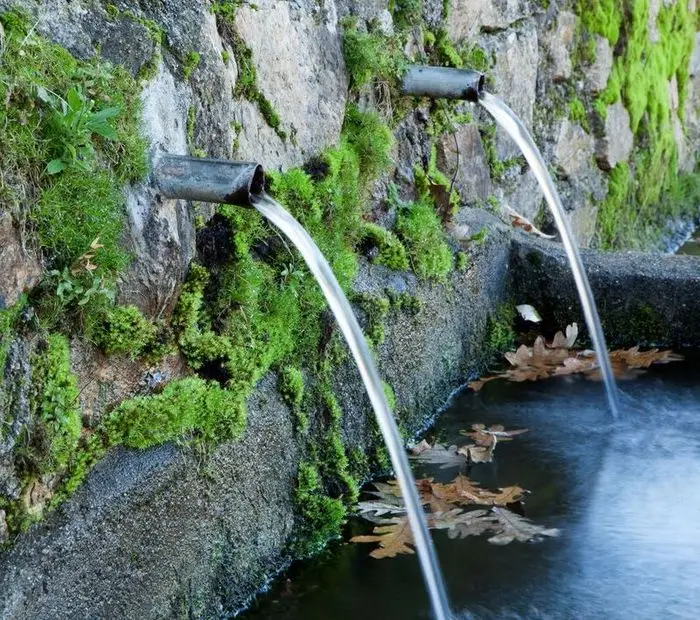 Das Bild zeigt zwei Metallrohre, die aus einer mit Moos bewachsenen Steinwand herausragen. Aus den Rohren fliesst Wasser in ein kleines Becken. Die Szene wirkt harmonisch, da die Natur mit den vom Menschen geschaffenen Elementen verschmilzt.