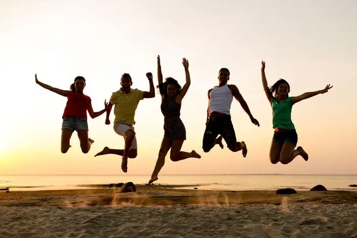 Das Bild zeigt fünf Personen, die in Eintracht gegen einen atemberaubenden Sonnenuntergang an einem ruhigen Strand jubelnd in die Luft springen und einen Moment der Freiheit und Begeisterung einfangen.